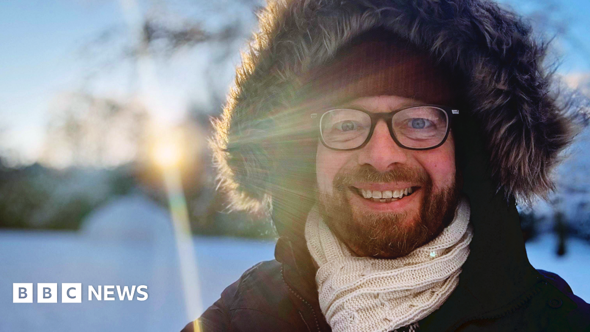 BBC reporter Ken Banks in snow, smiling at camera, wearing a thick hooded jacket with a fur trim round the hood and a scarf around his neck. The sun is shining in the background.