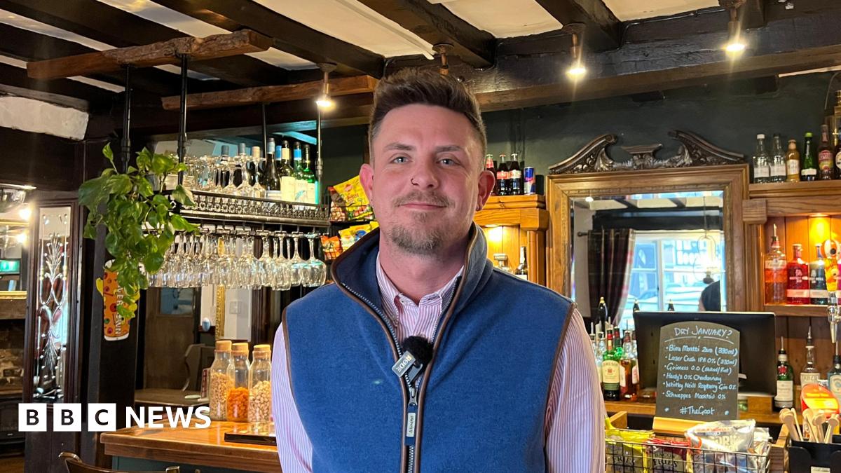 Ian Hillman wears a blue gilet and stands in front of the bar at his pub, a Sixteenth Century inn. He has a goatee, and is smiling.