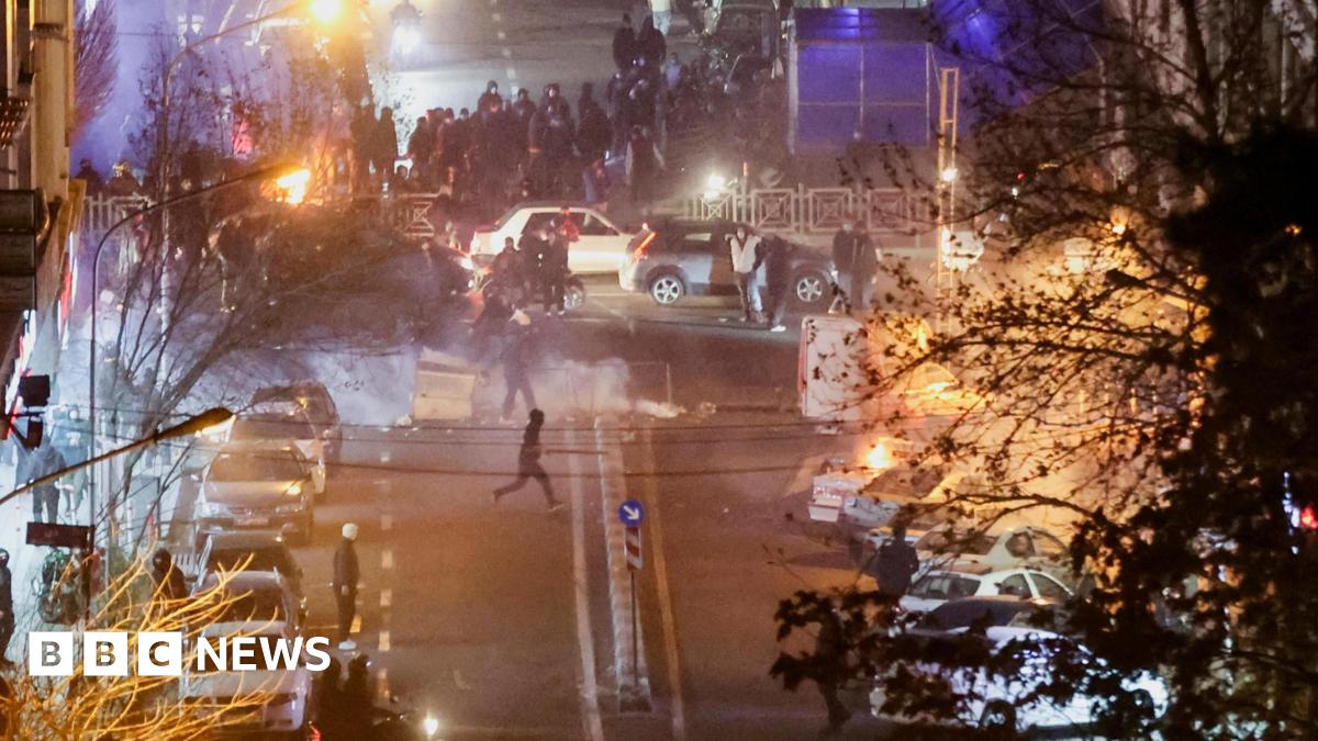 A street scene at night showing protesters gathering around parked cars. Smoke can be seen and there are some fires near the crowd.