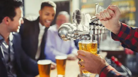 Getty Images Close up of a pint being pulled from a tap. In the background there are blurred images of young men in shirts and jackets sitting at the bar with full pints in front of them. They look cheerful.