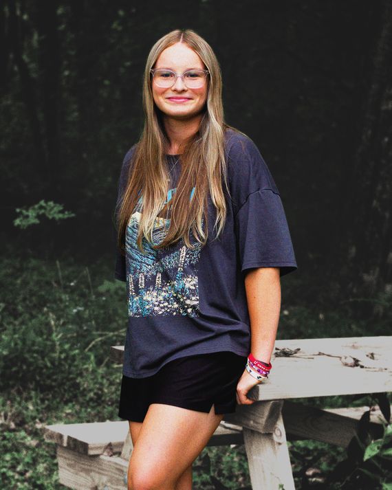 A girl with long blonde hair wearing glasses poses for a portrait in front of a picnic table.