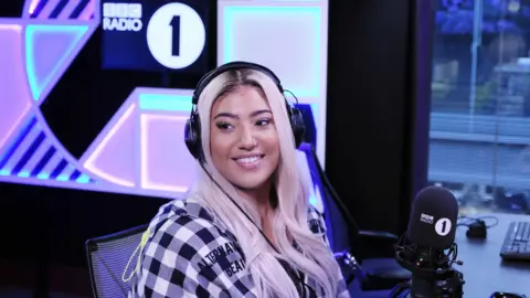BBC Tiffany Calver is sitting in a studio in front of a BBC Radio 1 microphone. She has headphones on is wearing a black-and-white checked jacket and has blonde hair.  