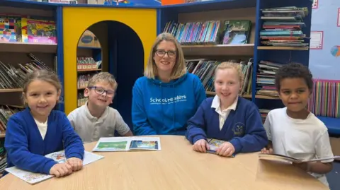 BBC / Grace McGrory The photo shows four schoolchildren, and one woman smiling at the camera. The pupils are wearing school uniform, with a boy and girl sitting on either side of the adult. The woman in the middle is wearing a bright blue hoodie with the word 'Schoolreaders' written on it. They are all sitting at a table with books in front of them. There are bookshelves filled with books behind them. 