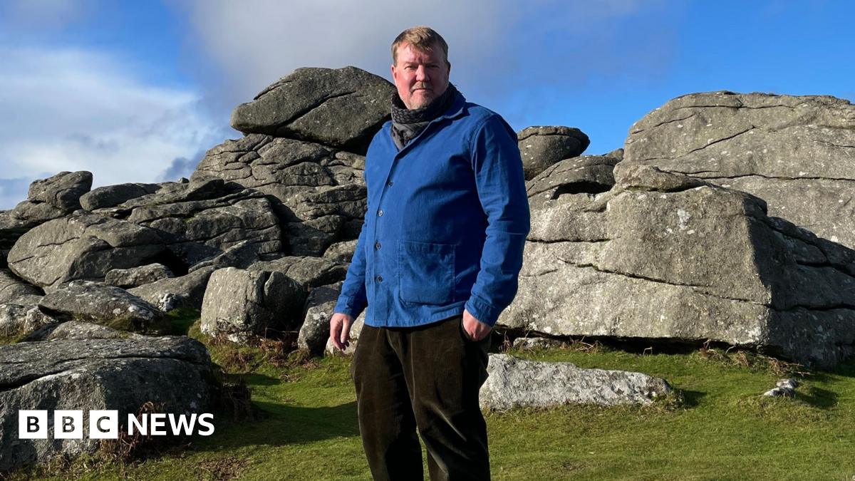 Artist and author, Alex Murdin, is wearing a blue jacket, and stands in front of Bonehill Rocks on Dartmoor. The grass is bright green and the sky is blue with some light clouds.