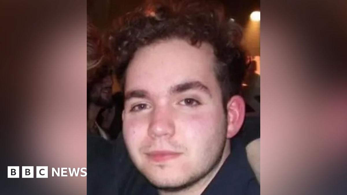 Head and shoulders photograph of Connor. He has dark wavy hair and is standing in a dark room with a spotlight in the background.