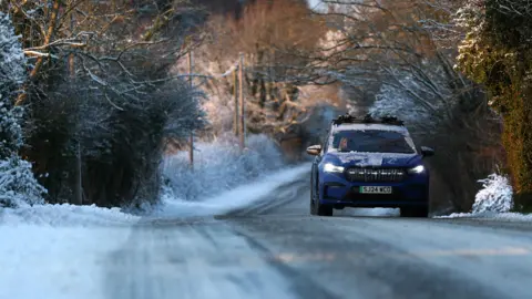 EPA A car drives along a snow-covered road in Mold, Wales. There is snow on the hedges surround the road, which is partially-obscured by snow and ice.