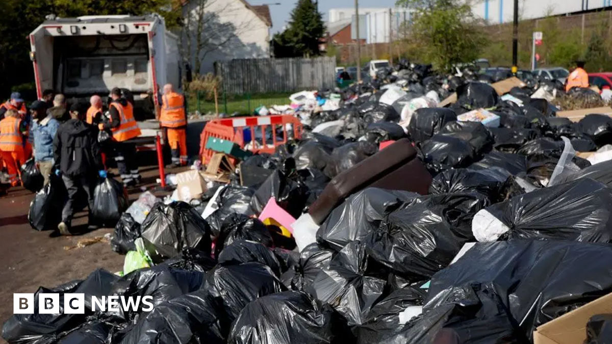 Dozens of bin bags are piled on top of one another on the right of the image with a bin lorry and people stood near it on the left hand-side of the image. Works with fluorescent jacket can also be seen in the image.