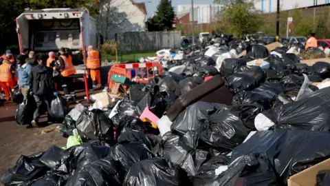 EPA Dozens of bin bags are piled on top of one another on the right of the image with a bin lorry and people stood near it on the left hand-side of the image. Works with fluorescent jacket can also be seen in the image.