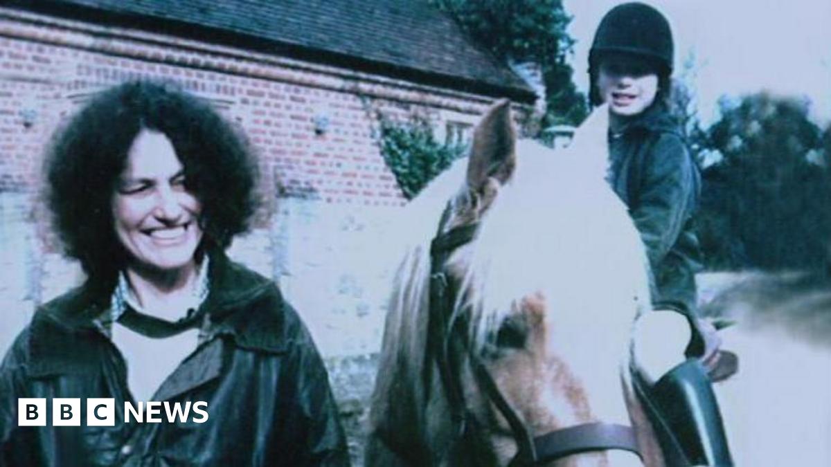 A woman with shoulder length dark curly hair and wearing a dark coloured jacket and light coloured blouse is smiling as she stands next to a young girl riding a horse. The girl is also smiling. A brick building and trees can be seen behind them.