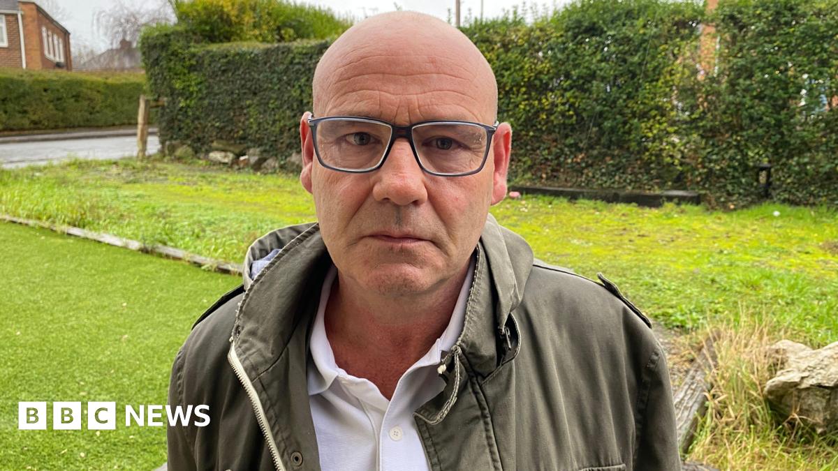 Barry Froggatt in a picture. He is standing on a front drive looking at the camera. He is making a serious face. He is a bald man wearing glasses, a white polo top and a green jacket.