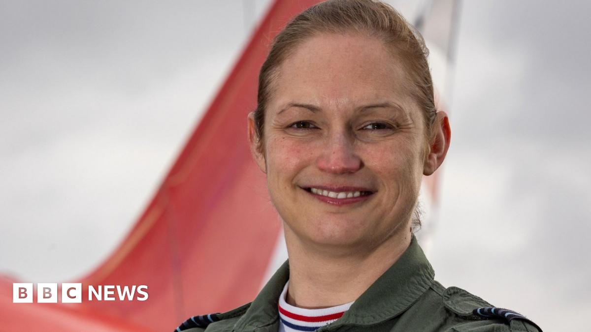 Head and shoulders shot of a smiling Wing Cdr Sasha Nash. She has fair hair which is tied back and is wearing overalls. There is the tail section of a Red Arrows jet in the background.