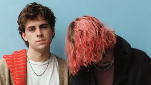 Royel Otis Press photo of Royel Otis standing against a plain blue background. Guitarist Royel Maddell covers his face with long, pink tousled hair