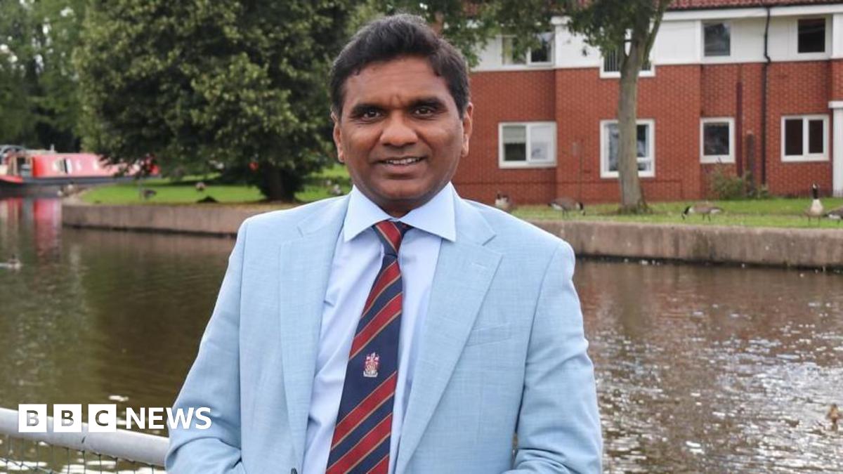 Dr Chandra Kanneganti wears a pale blue suit with a stripy blue and red tie. He is smiling, and posing next to the canal in Stoke-on-Trent, with a narrow boat in the background, along with waterside properties.