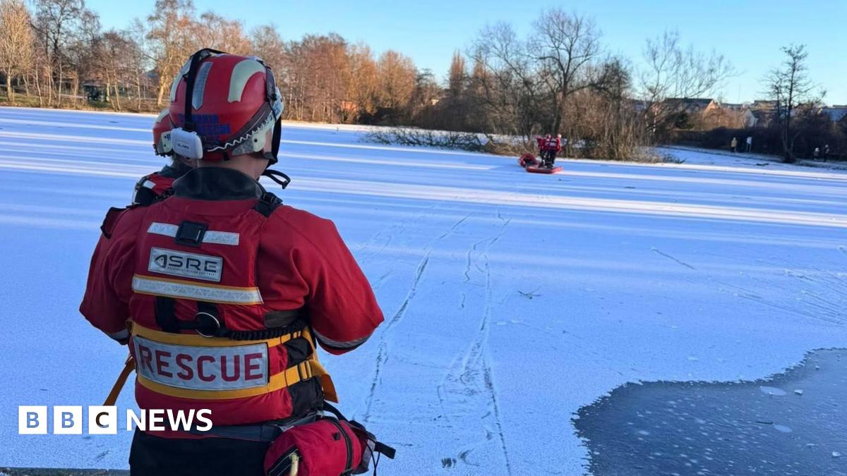 View of an iced over pond, with a light covering of snow. In the foreground is a member of the the fire service, wearing red jacket, marked "rescue" and a helmet and an ear piece. Further along the pond are members of Cumbria fire service. In the background trees are catching the winter sunlight.