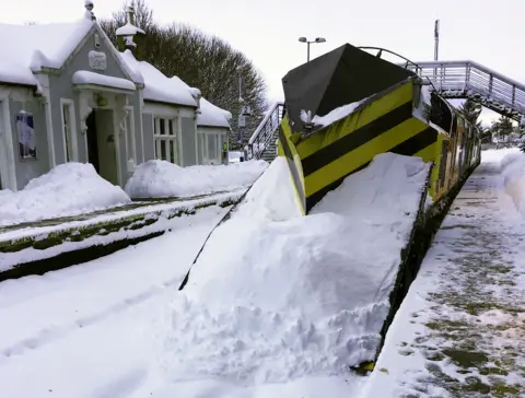 Network Rail A snowplough train covered in snow at a train station.