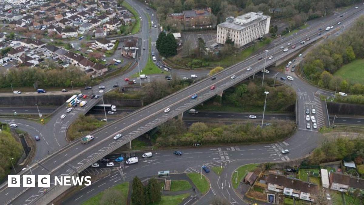 An ariel view of Manadon roundabout in Plymouth. There is a large area at the centre with trees either side of it. There are cars driving on the road and housing in the top left corner. There are two larger buildings to the right of the houses.
