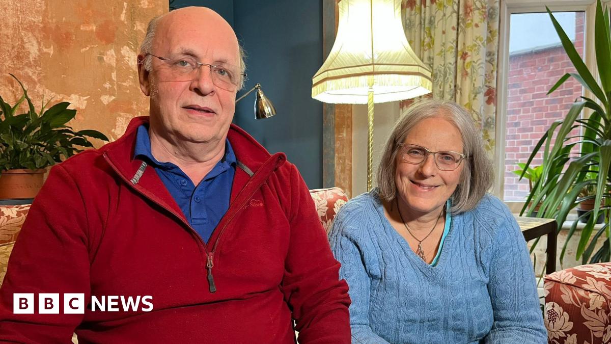 Millie Bobby Brown and David Harbour in character as Eleven and Jim Hopper. Eleven wears red shorts over grey leggings and a grey jumper. Jim Hopper wears a black leather jacket, a black beanie hat and dark trousers.They are speaking to director Frank Darabont on the set of Stranger Things. There is a vintage advertisement posted in the background with debrief on the ground.