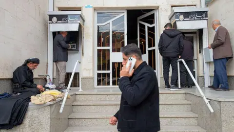 Mobina / Getty Images Men outside a bank using ATMs and one is walking by on his mobile phone.