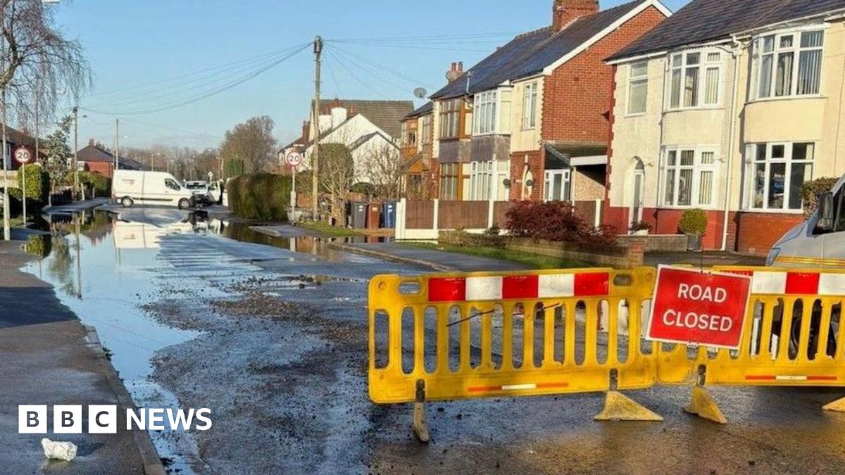 Image of water flooding sections of the road next to houses. Yellow barriers are placed in front with a red sign saying 'road closed'.