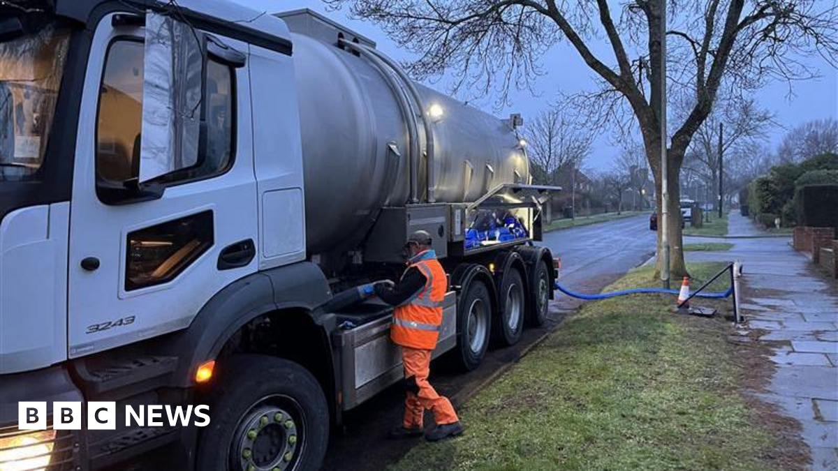 A man dressed in a high-vis suit stands near a tanker lorry which appears to be plugged into a water supply. It is early morning light.