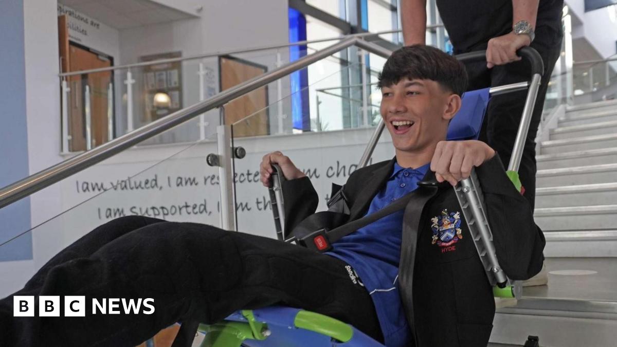 Lucas, who has brown hair, is wearing a school uniform and is smiling as he is lowered down the stairs of his school in an evacuation chair.