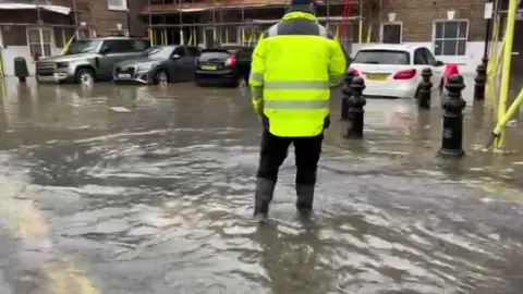 A man in a high vis jacket and waterproof boots stands in a car park which is submerged in a foot of water. 