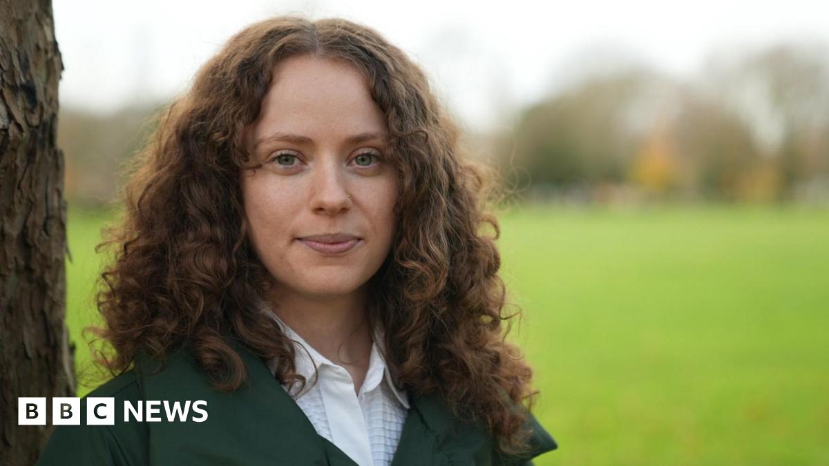 Holly stares to camera. She is in focus with dark, long curly hair and is leaning against a tree-trunk while the background is out of focus but looks like a green field.