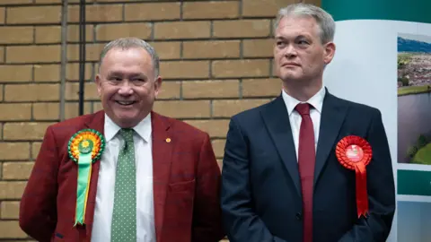 Getty Images Two men - one smiling with a green Plaid Cymru badge and one not smiling with a red Labour badge - stand on a stage next to each other.