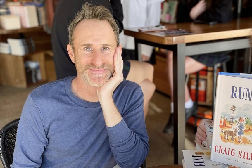 Craig wears a blue long sleeve top and rests his head in the palm of his hands as he smiles