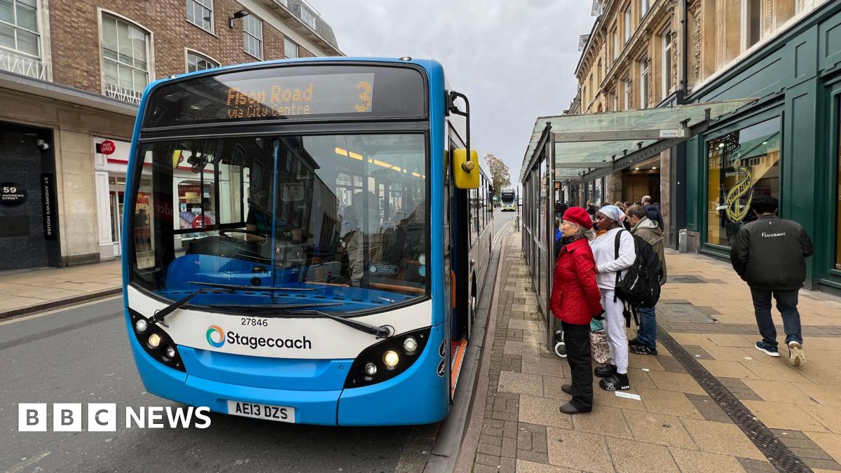 A blue number three Stagecoach bus in a street lined by shops, with passengers waiting to board underneath a bus shelter.