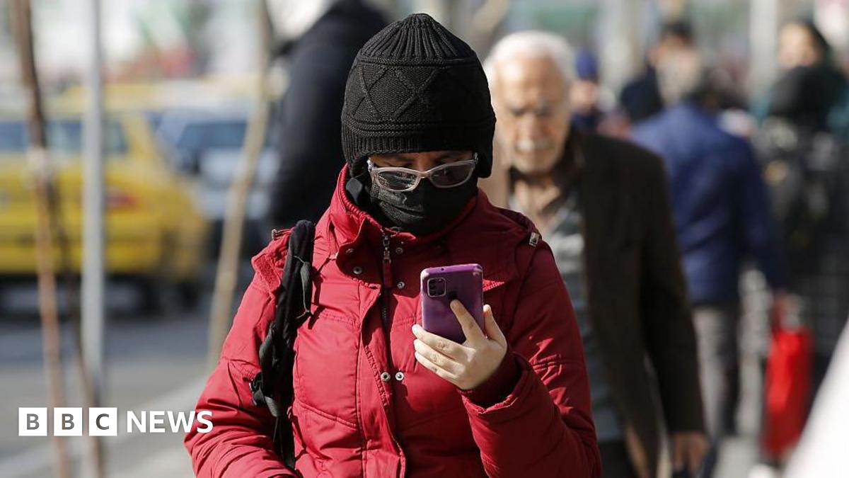 A person in Iran holding a smartphone. They are wearing a big red jacket, a black hat, a black face mask and glasses.