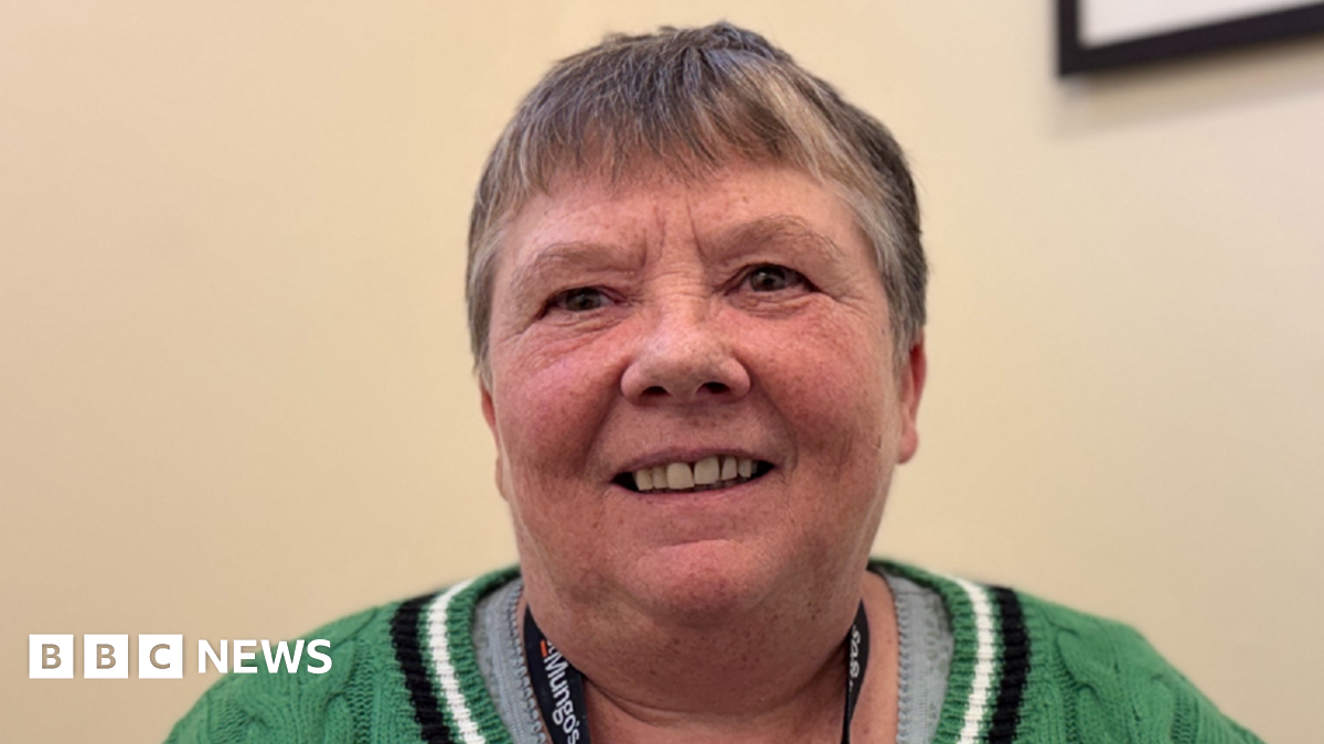 A picture of Wendy Dodds, who is wearing a green jumper and grey shirt and smiling. She has short, dark grey hair.