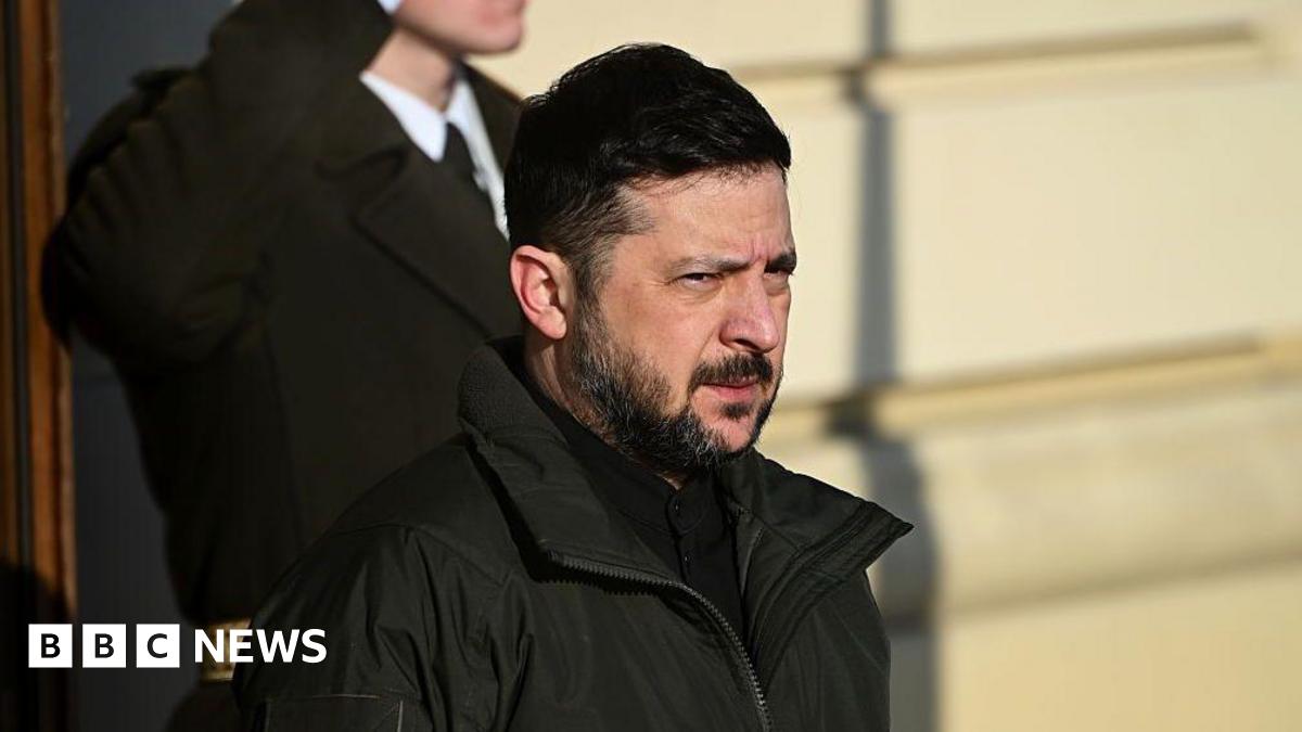 A man in a dark beard and black coat against a yellow background