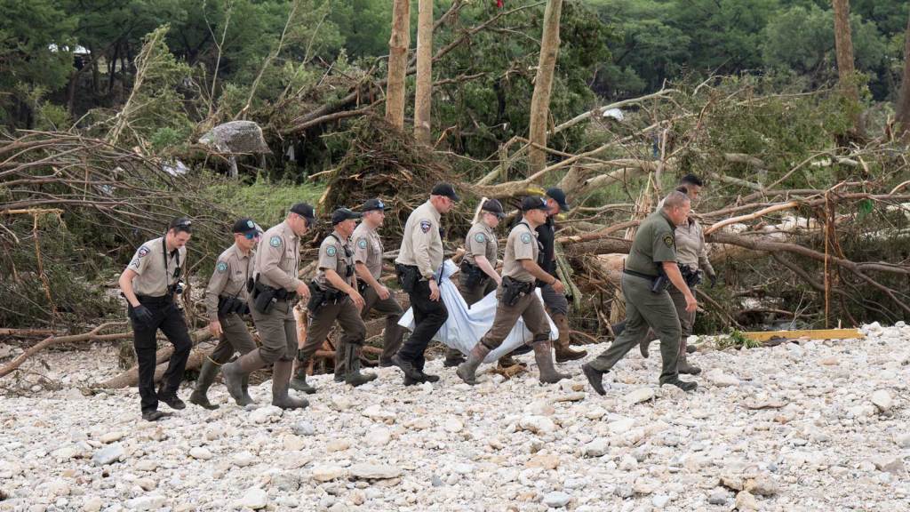 Photo of a group of uniformed men. Some are carrying the body of a flood victim, who is covered in a white body bag or sheet.