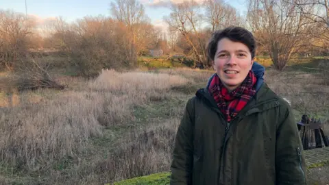 Kim Man with brown hair wearing a scarf and green jacket smiling in a park