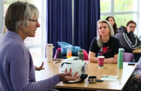 Joan Lyon, a 75-year-old woman, with grey bobbed hair and glasses, wearing a lilac cardigan stands with her arms outstretched. She is speaking while three students watch.