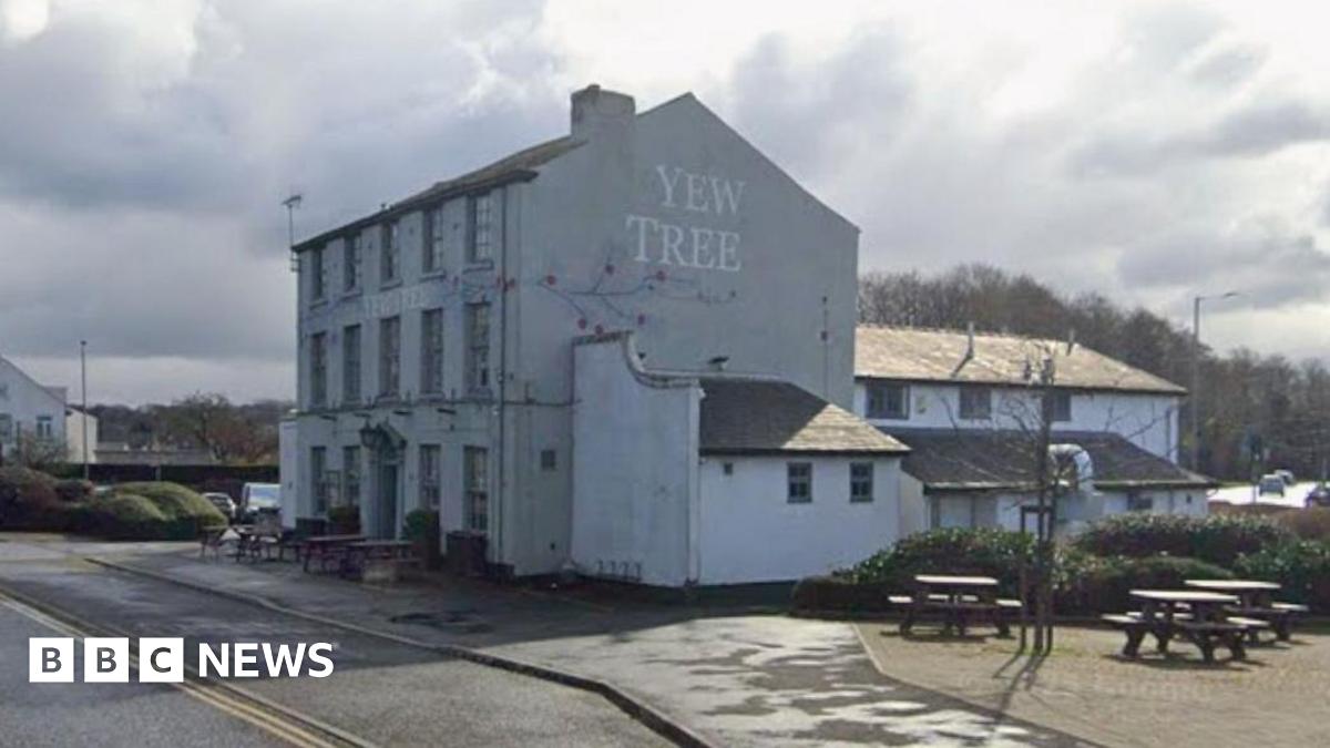 Three-storey old white building with the name Yew Tree emblazoned on the side. Wooden benches and tables surround it with a road in front.