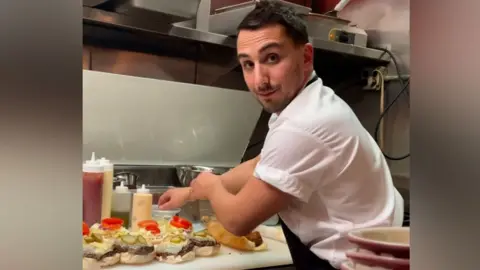 Seth Gerber Chef Seth Gerber wearing whites and preparing burgers in a professional kitchen and turning back to smile at the camera