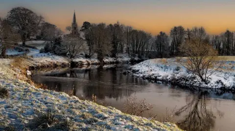 BBC WEATHER WATCHER - NIGEL KELLY A river with snow along the banks and a church and trees in the distance. The sky is orange.