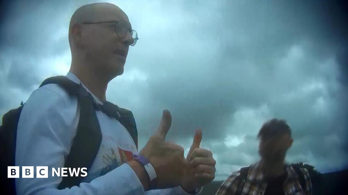 Mr Lawler, a bald man wearing a white long-sleeve t-shirt with a partially-visible graphic on the front, gives a double thumbs-up gesture towards somebody not in frame. He is wearing a rucksack and standing next to another man, whose face is blurred.