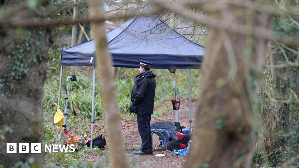 A police officer stands near a gazebo in a wooded area. The gazebo has lots of items under it on the ground.