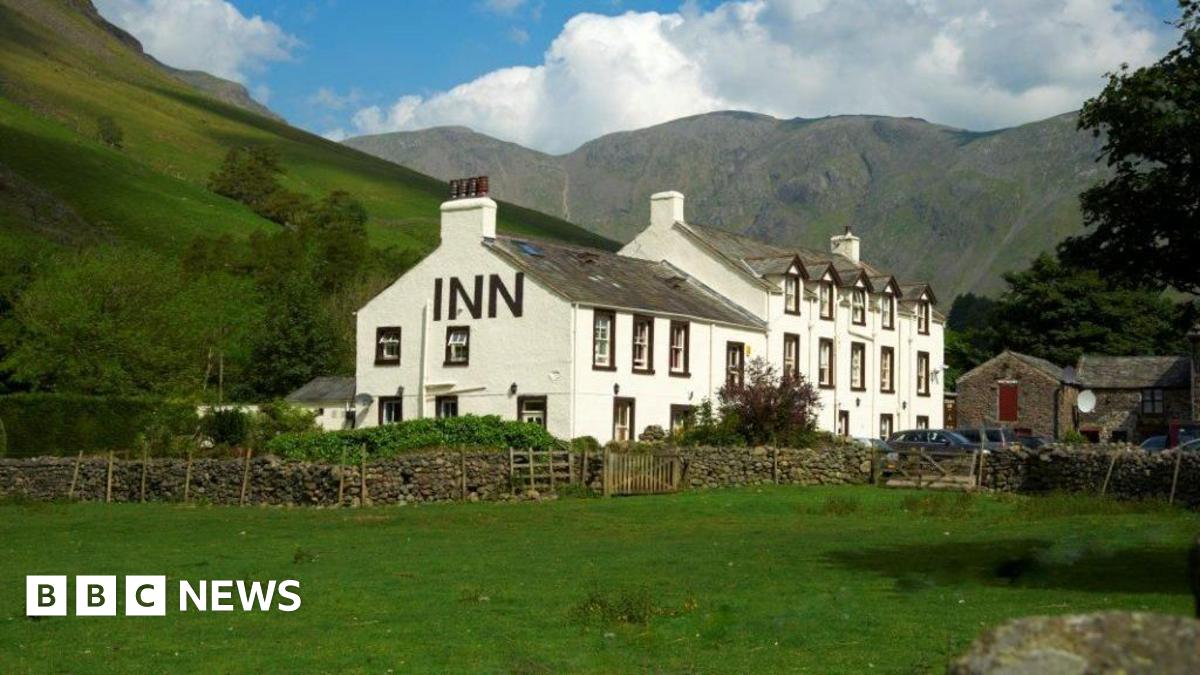 A general view of the Wasdale Head Inn. It is a traditional Lakes building, painted white with black detailing around the sash windows. and a slate pitched roof. It is set within a remote setting, with the mountains behind it and a grassy field in front of it