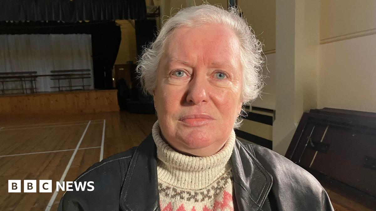People are sitting and a crowd is standing. A man is holding a wreath. Some are holding roses.