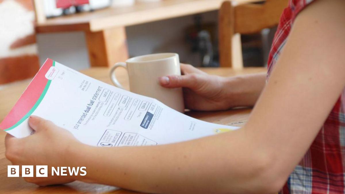 A woman wearing a checked top sits at a  wooden table, holding a bill in one hand and a mug in the other.