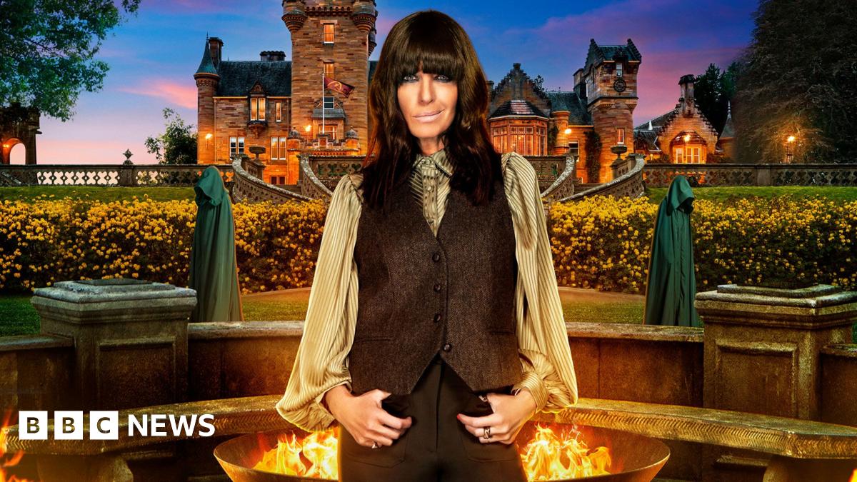 Claudia Winkleman stands in front of a firepit and behind that the Traitors castle in the distance and smiles at the camera. Two hooded Traitors are visible at either side of Claudia in the background