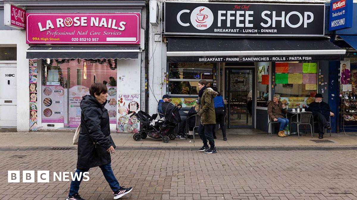 A woman in a black coat walks along a high street in Walthamstow, passed a Coffee shop and a nail salon with bright pink signage.