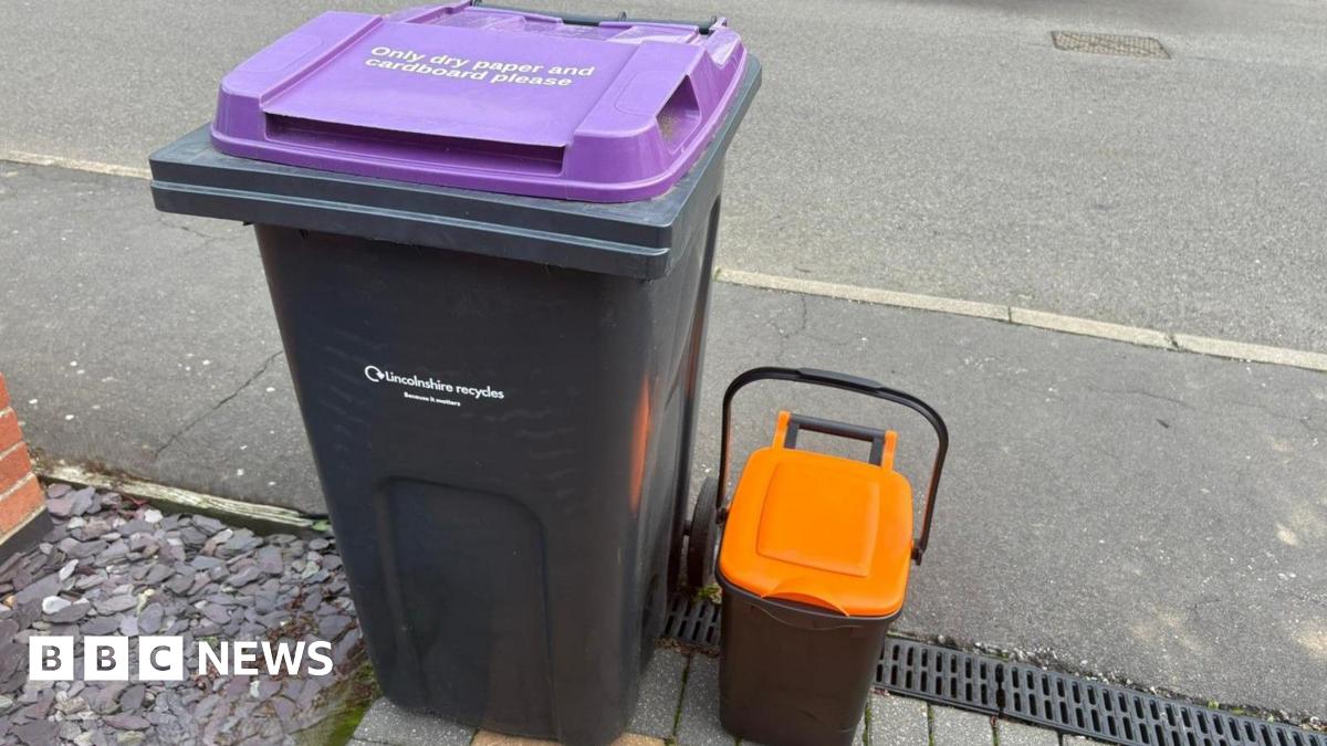 A black bin that says 'Lincolnshire recycles' on it in small white font with a purple lid that says 'Only dry paper and cardboard please'. Next to it is a small black caddy with a purple lid. It is situated at the end of a driveway next to a grey footpath and road.