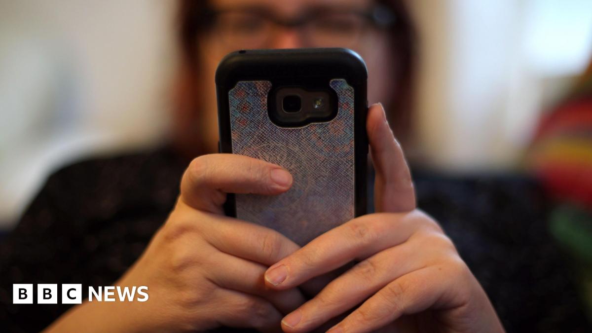 A blurred person wearing glasses holds up a mobile phone with both hands.