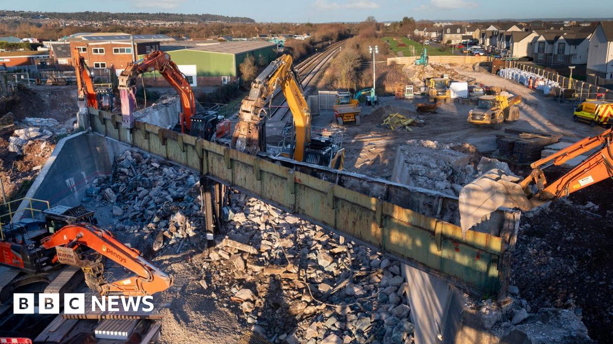 Diggers and other construction machinery are surrounded by rubble as a bridge is demolished over a railway track. It is a sunny winters day in a residential area.