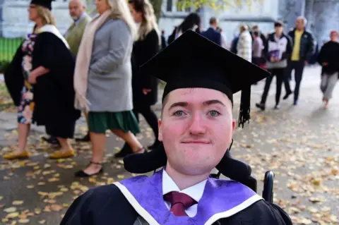 Ben Morris is shown on graduation day wearing a traditional black and purple gown with a graduation hat.